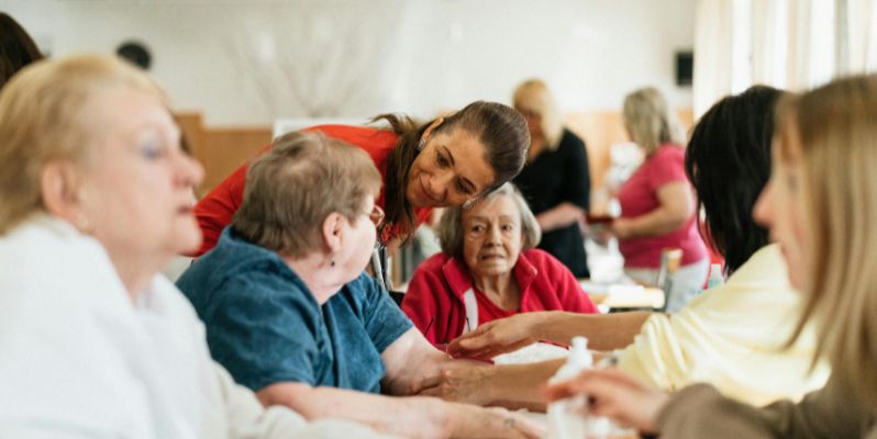 aged care worker leaning over to talk to a group of elderly women sitting around a table