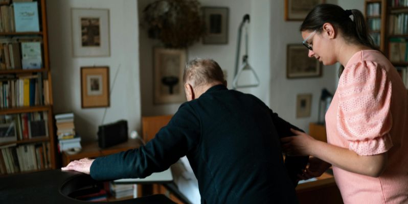 Women helping an elderly man stand up from a chair